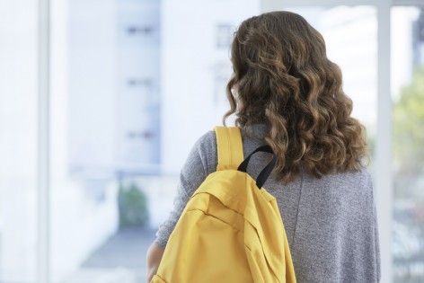 Mädchen, ca 14 Jahre, steht mit dem Rücken zur Kamera in einem Schulflur und schaut aus dem Fenster auf den Schulhof.