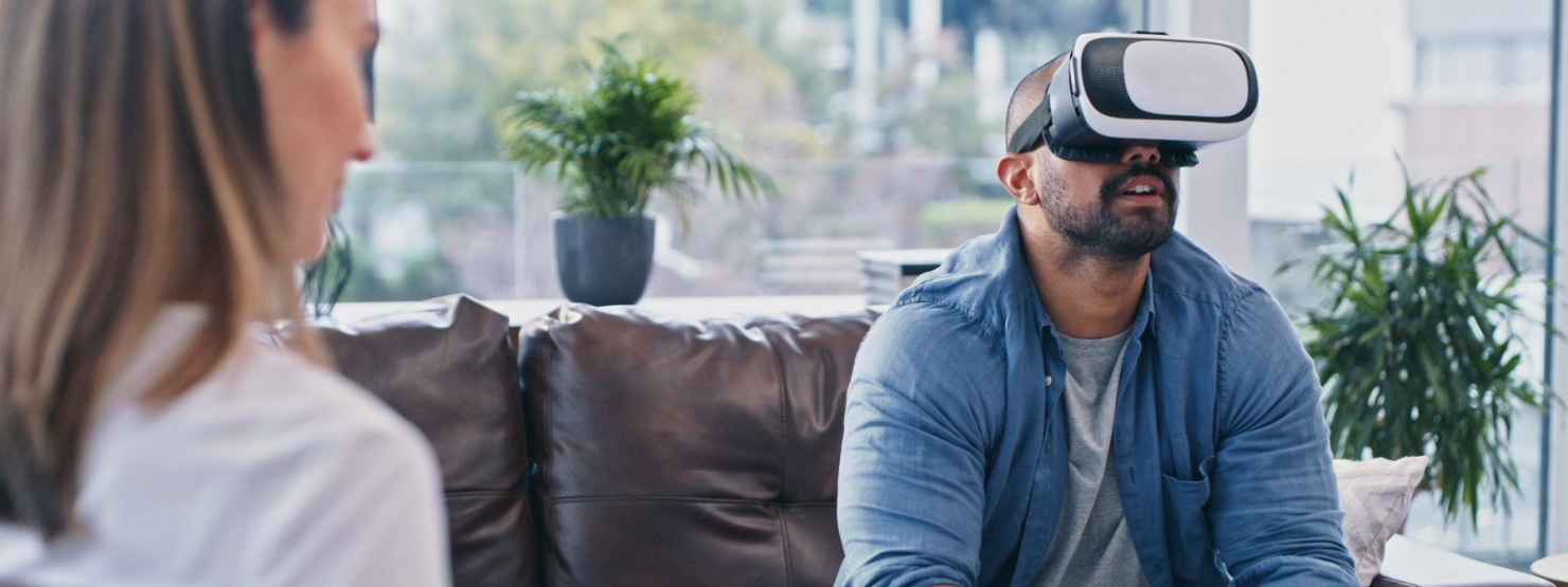 Ein etwa 35 jähriger Mann sitzt bei einer Therapeutin. Dabei trägt er eine Virtual Reality.