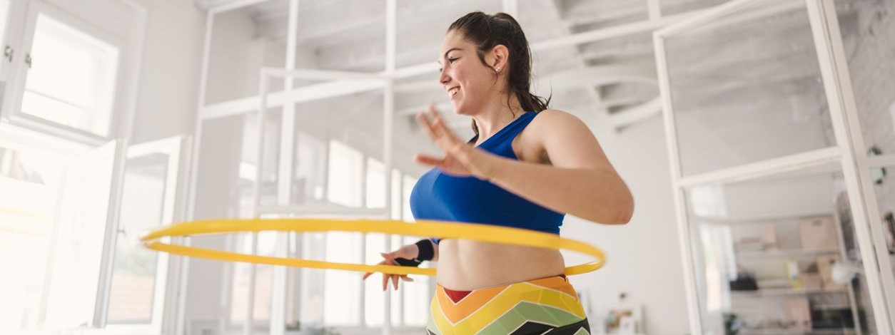 Ein Seniorenpaar steht auf einer Wiese und macht Sport mit einem Hula Hoop. Beide lachen.