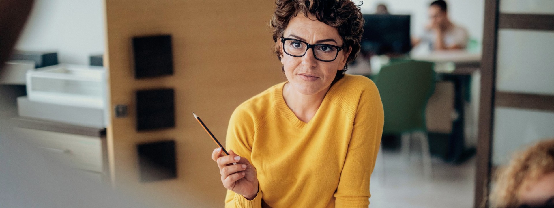 Eine etwa 40 jährige Frau in einem gelben Pullover und mit braunen Locken steht an einem Schreibtisch im Büro. Sie erklährt ihren Kollegen etwas und hebt dabei die Hand. Sie hält einen Stift in dieser Hand.