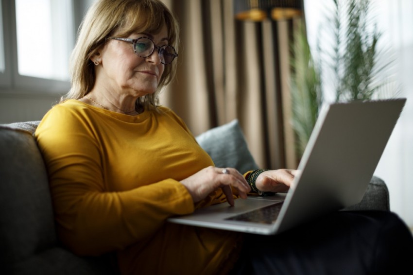 Eine Seniorin sitzt mit einem Laptop auf dem Schoß auf einem Sofa.