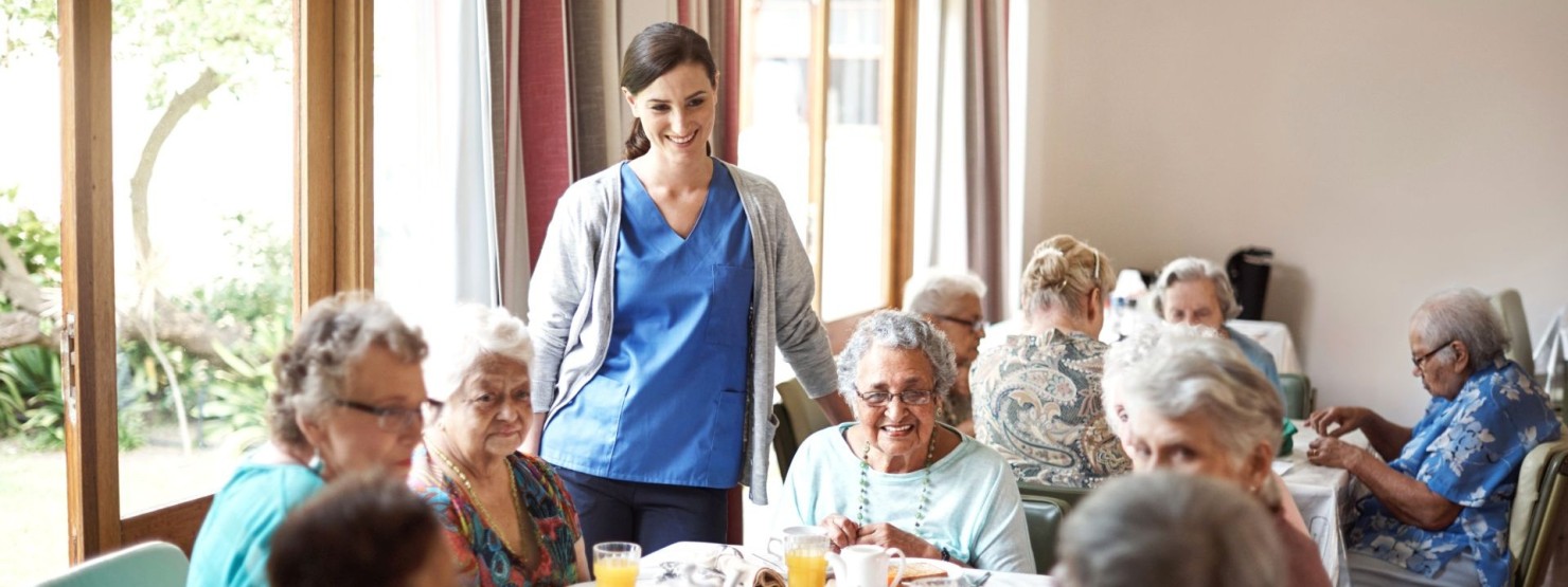 Eine Gruppe Seniorinnen sitzen zusammen am Tisch im Seniorenheim und frühstücken. Eine Pflegekraft steht neben dem Tisch.