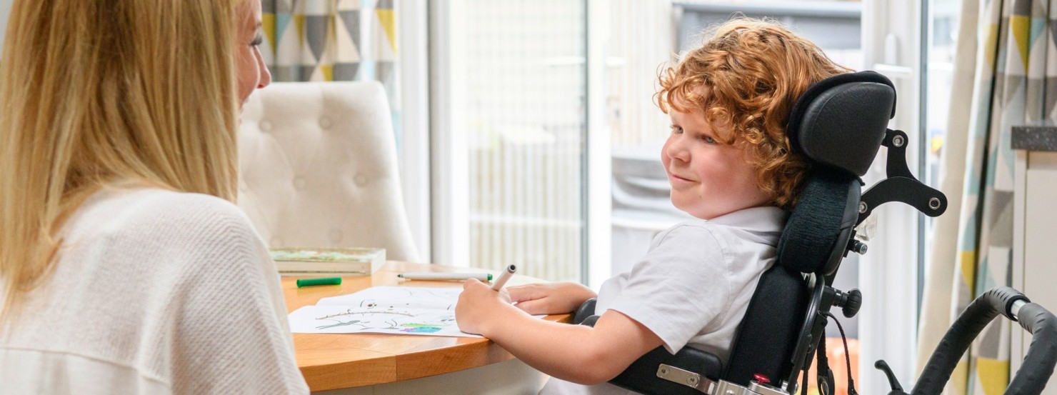 Ein etwa 10 jähriger Junge sitzt in einem Rollstuhl an einem Tisch und malt auf ein Blatt Papier. Ihm sitzt eine Frau mit einer Katze auf dem Schoß gegenüber.