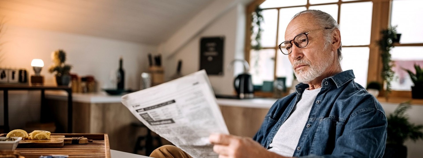 Ein etwa 60 jähriger Mann sitzt an einem Tisch und liest eine Zeitung. Er trägt eine Brille.