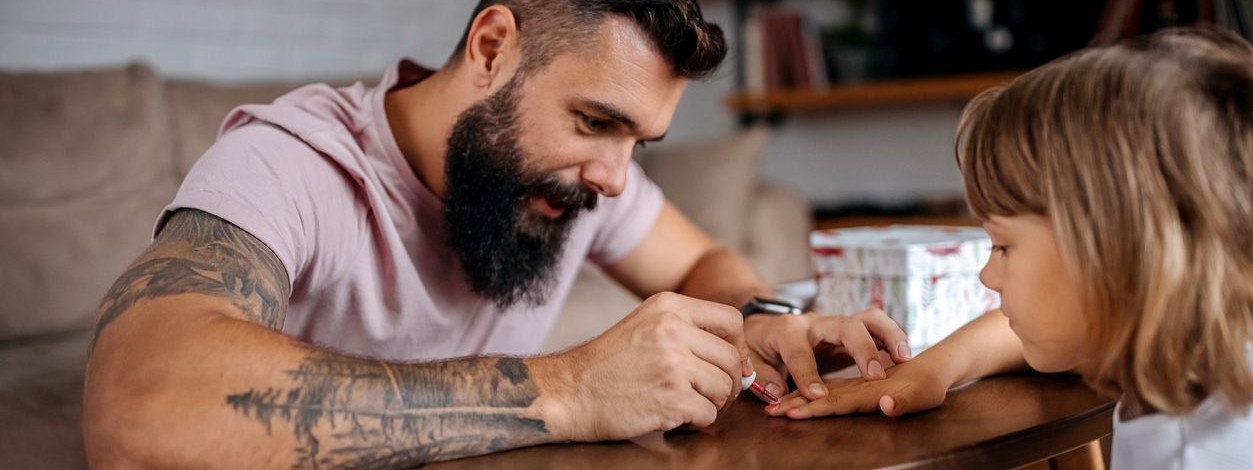 Ein etwa 30 jähriger Mann sitzt mit seiner Tochter gemeinsam am Wohnzimmertisch. Er bemalt ihre Fingernägel mit Nagellack für Kinder. Seinen Arm zieren mehrere Tattoos. 