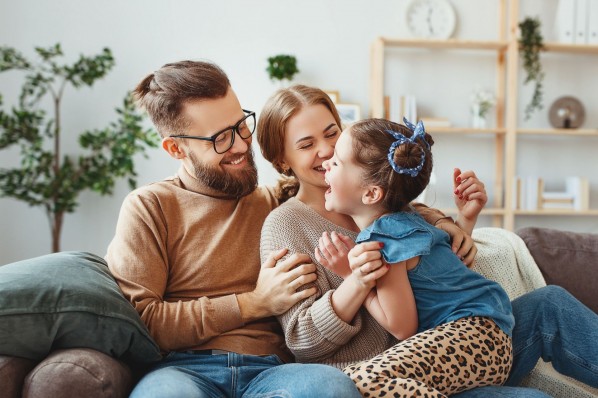 Eine Familie sitzt auf einem Sofa in einem Wohnzimmer. Alle drei lachen ausgelassen und haben Spaß.