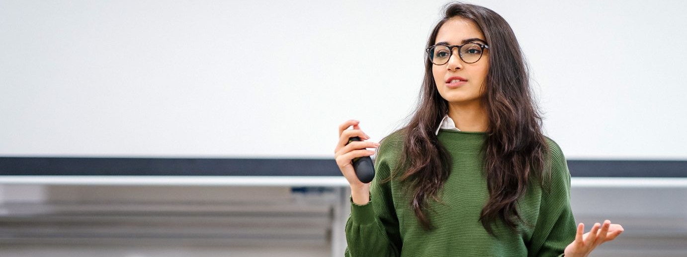 Eine etwa 25 jährige Frau steht in einem Hörsaal und referiert vor einer Leinwand. In der rechten Hand hält sie die Fernbedienung für den Beamer.