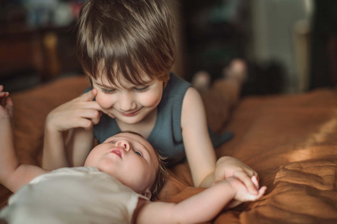 Ein Baby und sein 3 Jahre alter Bruder liegen zusammen auf dem Bett. Das Baby liegt auf dem Rücken. Der Bruder liegt auf dem Bauch. Sie schauen sich an und halten Händchen.