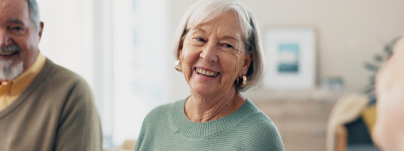Zwei Seniorinnen sitzen auf einer Couch im Wohnzimmer und trinken gemeinsam Kaffee. Beide sind zueinander gedreht und lächeln sich an.