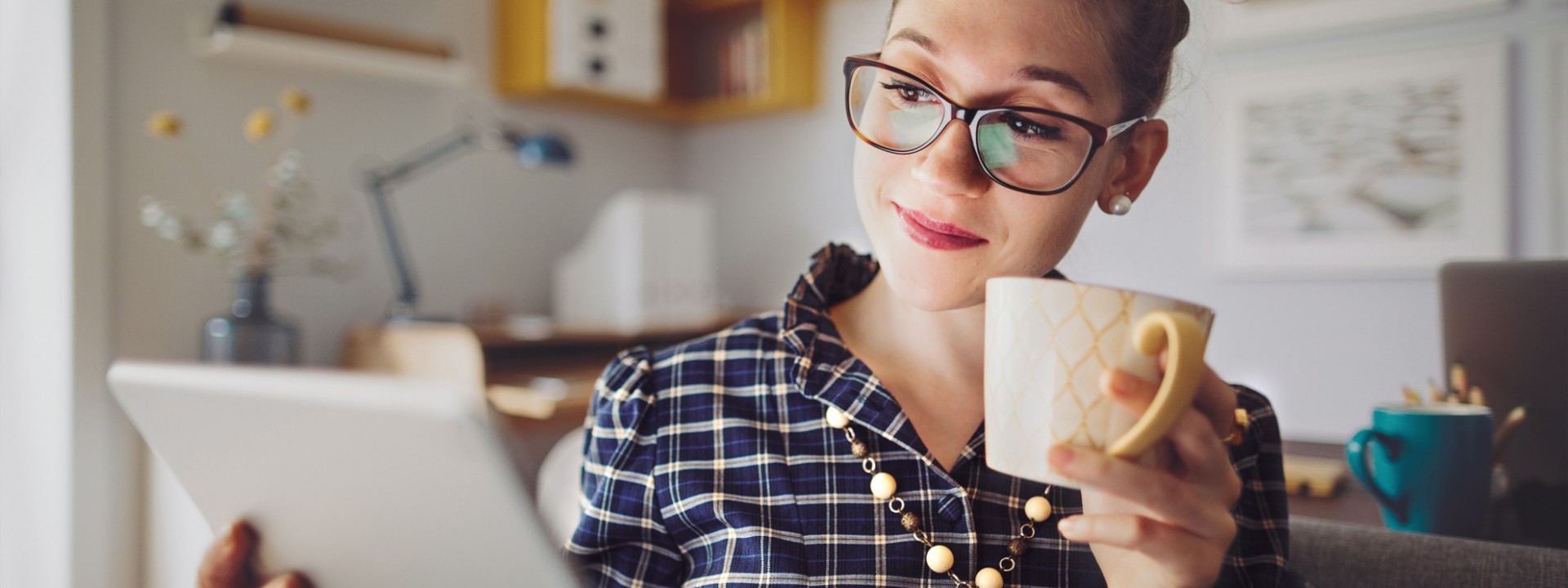 Eine etwa 30 jährige Frau befindet sich zu Hause. Sie hält in der einen Hand ein Tablett, auf welches sie schaut, und in der anderen Hand eine Tasse.