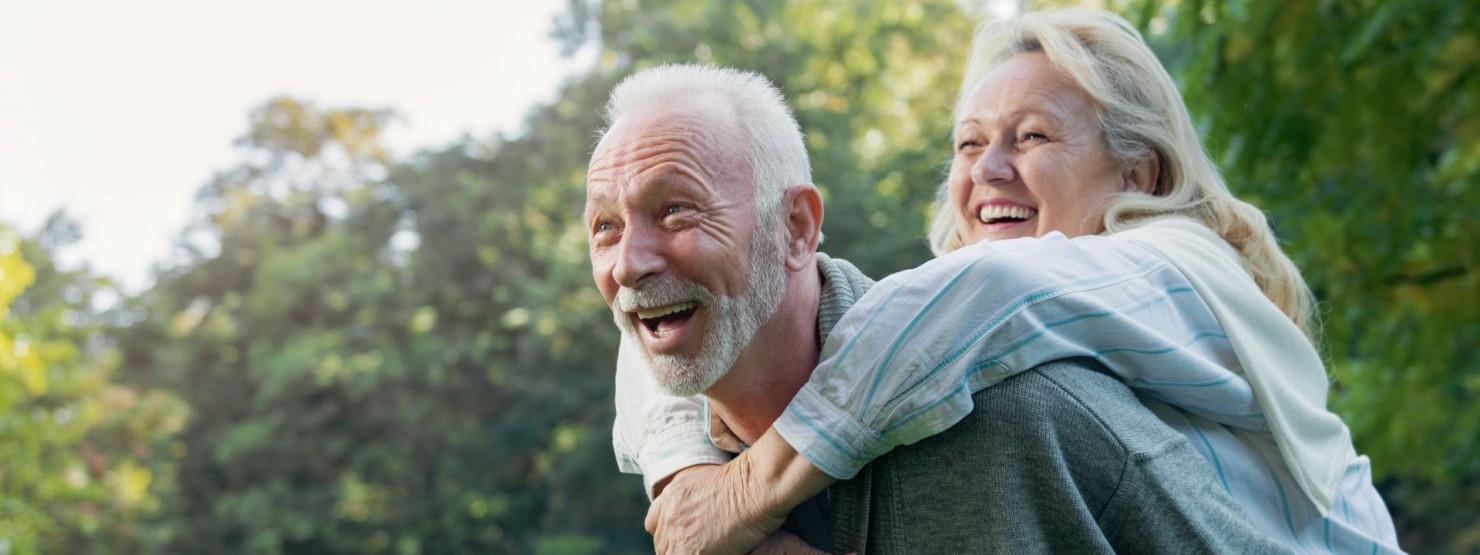 Ein Seniorenpaar hat gemeinsam in der Natur Spaß. Der Mann trägt die Frau auf dem Rücken. Beide lachen ausgiebig.