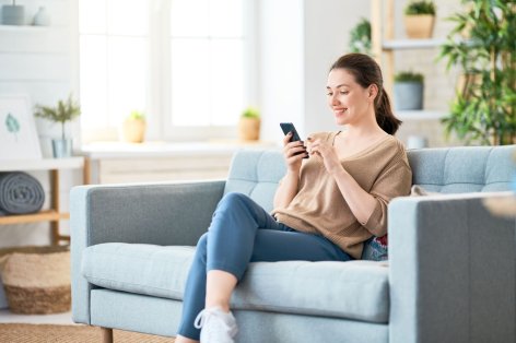 Eine etwas 30 jährige Frau sitzt auf der Couch hat ein Handy in der Hand. Sie lächelt.