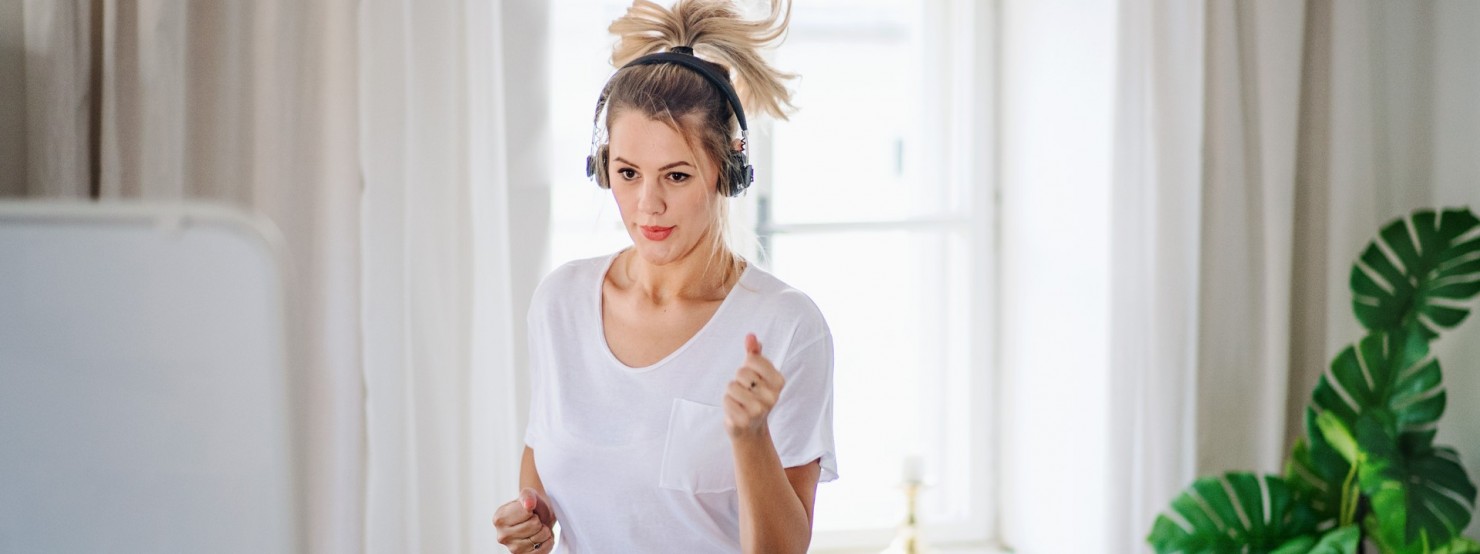Eine junge Frau steht im heimischen Wohnzimmer und treibt Sport. Sie hört dabei Musik mit Kopfhörern.