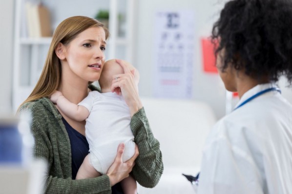 Eine junge Mutter steht mit Ihrem Baby auf dem Arm einer Ärztin gegenüber. Sie sieht ängstlich aus.