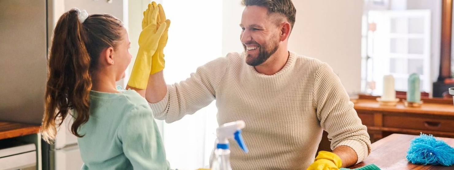 Eine etwa 30 jährige Frau hockt auf dem Boden im Wohnzimmer. Neben ihr am Couchtisch steht ihre etwa 6 jährige Tochter. Beide tragen Gummihandschuhe und putzen den Tisch. Um beide herum stehen Reinigungsmittel und Putzeimer.