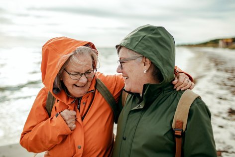 Zwei Seniorinnen spazieren lachend am Meer entlang.