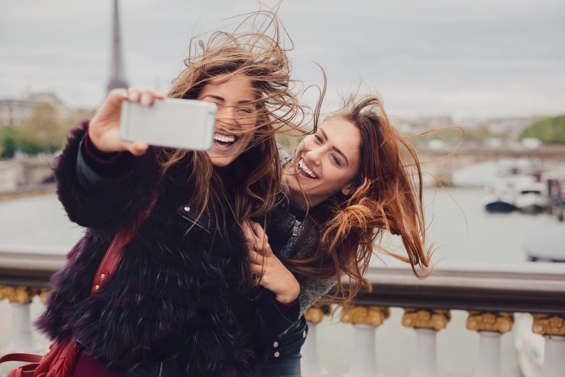 Zwei etwa 18 Jahre alte Frauen mit langen Haaren stehen auf einer Brücke und machen ein Selfie. Sie lachen.