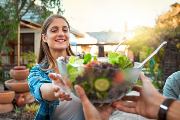 Freunde grillen im Garten. Eine junge Frau bekommt von einem Mann die Salatschüssel gereicht.