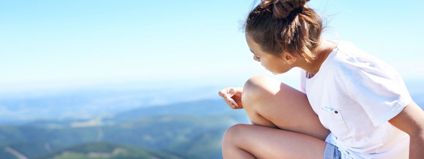 Eine etwa 20 jährige Frau sitzt auf einem Hügel in der Natur und verarztet ihr Knie mit einem Pflaster.