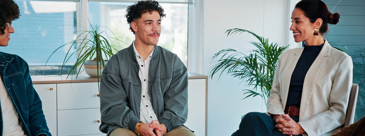 Ein etwa 25-jähriger Mann sitzt gemeinsam drei anderen Personen in einer Praxis zusammen. Alle Personen nehmen an einer Gruppentherapie teil und schauen sich an.