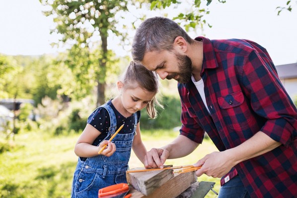 Ein etwa 5-jähriges Mädchen und ihr Vater stehen im Garten und zeichnen eine Linie auf ein Holzstück.