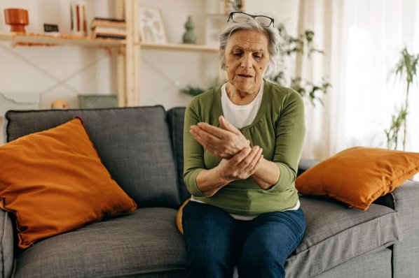 Eine Seniorin sitzt auf der Couch und hält ihr Handgelenk besorgt fest.