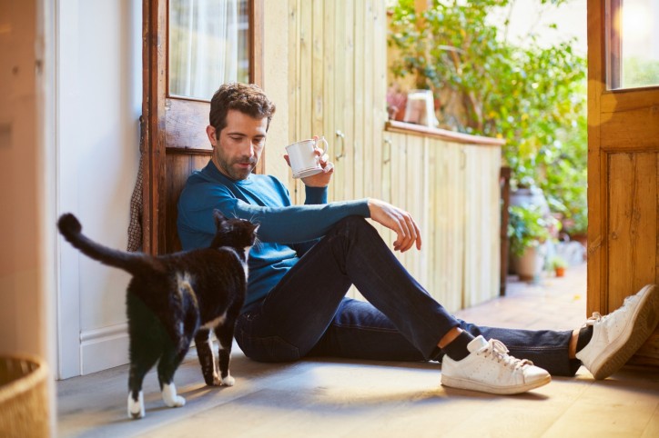 Ein Mann sitzt in der offnen Tür mit einer Kaffeetasse in der Hand. Seine Katze kommt um die Ecke.