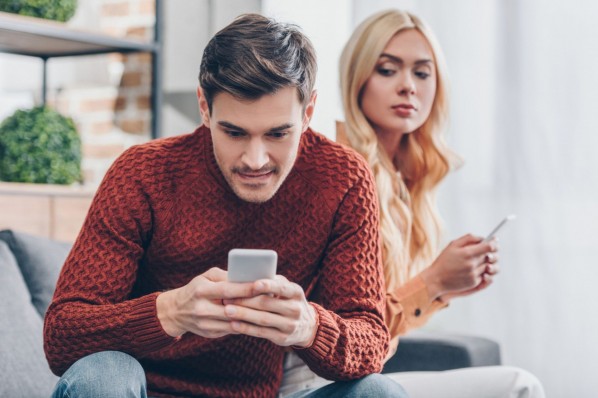 Eine etwa 30-Jährige schaut ihrem Freund dabei zu wie er auf sein Handy schaut. Sie sitzen auf einem Sofa.
