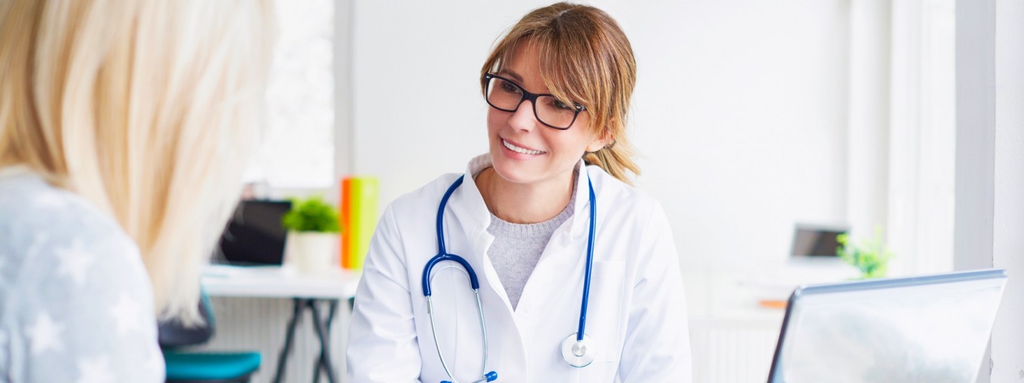 Eine Ärztin sitzt in einem Behandlungszimmer vor ihrem Laptop und berät eine Patientin.