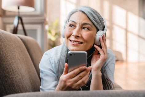 Eine Seniorin liegt auf einem Sofa und hört über Kopfhörer den AOK-Podcast "ausgesprochen menschlich - Selbsthilfe auf Sendung".