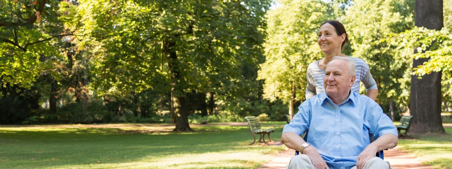 Eine Frau schiebt einen Senior im Rollstuhl durch den Park.