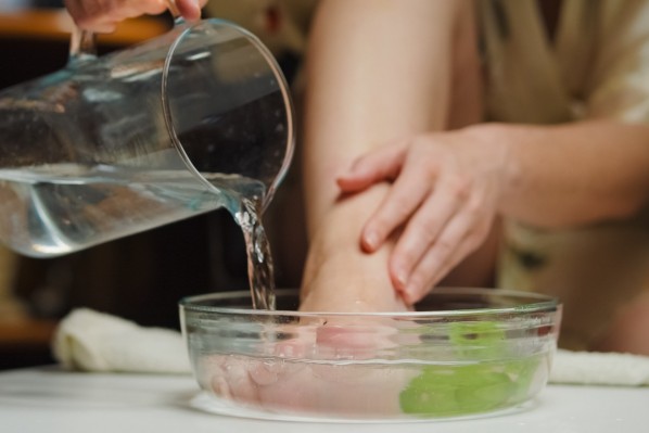 Eine Frau macht ein Fußbad. Ihr rechter Fuß steht in einer Glasschüssel und sie gießt Wasser zu.