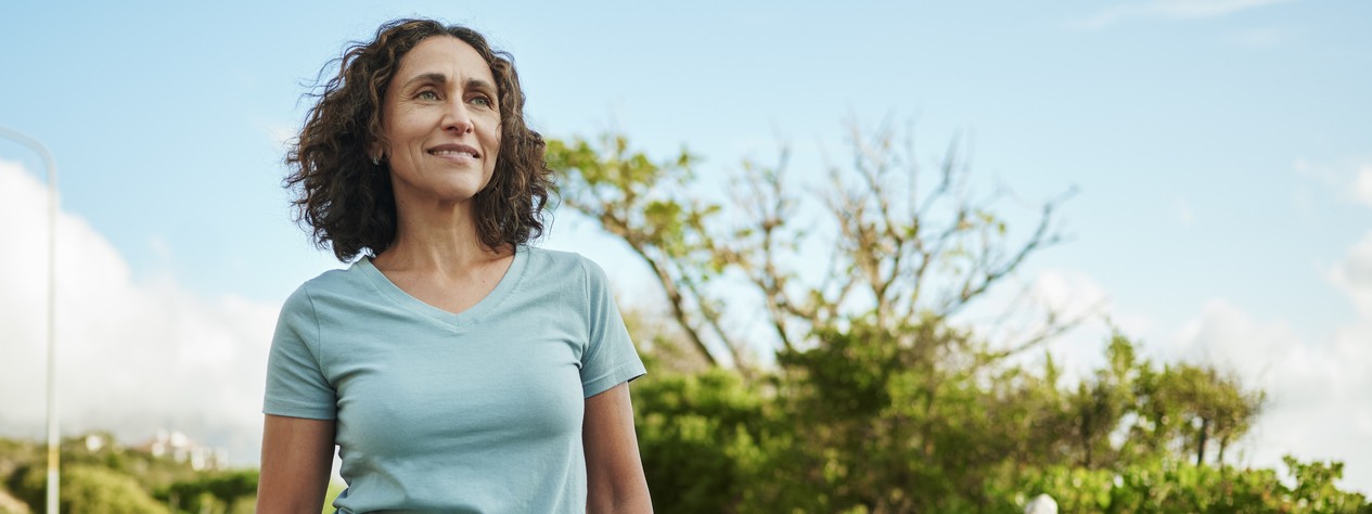 Eine etwa 40 jährige Frau mit Sonnenbrille und Sonnenhut sonnt sich am Strand. Dabei lächelt sie und schaut in die Ferne.