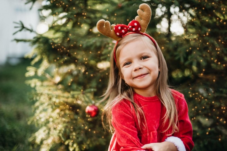 Ein etwa 4 jähriges Mädchen steht vor einem geschmückten Weihnachtsbaum und lächelt.