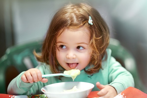 Ein etwa 3-jähriges Mädchen löffelt Kartoffelbrei aus einer Schüssel. 