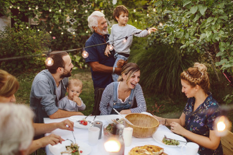 Mehrere Generationen einer Familie befinden sich zu einem sommerlichen Essen draußen in einem Garten. Der Großvater, etwa 65 Jahre, hat seinen Enkelsohn, etwa 4 Jahre, auf dem Arm und inspiziert mit ihm die zwischen den Bäumen gespannte Lichterkette. Zwei Frauen und ein Mann im Alter von etwa 30 bis 40 sitzen am Tisch in entspannter Atmosphäre beim Essen und Unterhalten. Der Mann hat ein weiteres, etwa zweijähriges Kleinkind auf dem Schoß und blickt zu Großvater und Kind hinüber. 