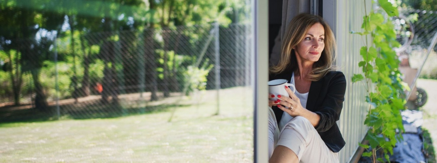 Eine etwa 40 jährige Frau lehnt sitzend in einem Türrahmen zur Terrasse und schaut in den Garten. In der Hand hält sie eine Tasse.