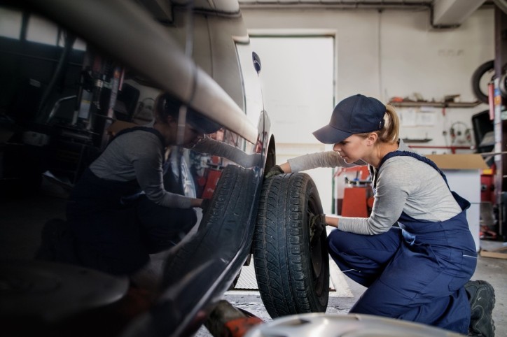 Eine etwa 20 Jahre alte Frau montiert in einer Werkstand einen Reifen an ein Auto. Sie kniet neben dem Auto und hält den Reifen mit beiden Händen fest.