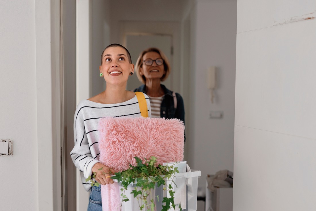Ein etwa 20-jährige Frau kommt in ein Zimmer und schaut glücklich lächelnd nach oben in den Raum. Sie trägt einen Plastebehälter mit einer Pflanze, einem Kissen und anderen Dingen. Hinter ihr sieht man den Flur einer leeren, hellen Wohnung. Ihr folgt ihre Mutter, etwa Mitte 40, die ebenso glücklich in den Raum blickt.