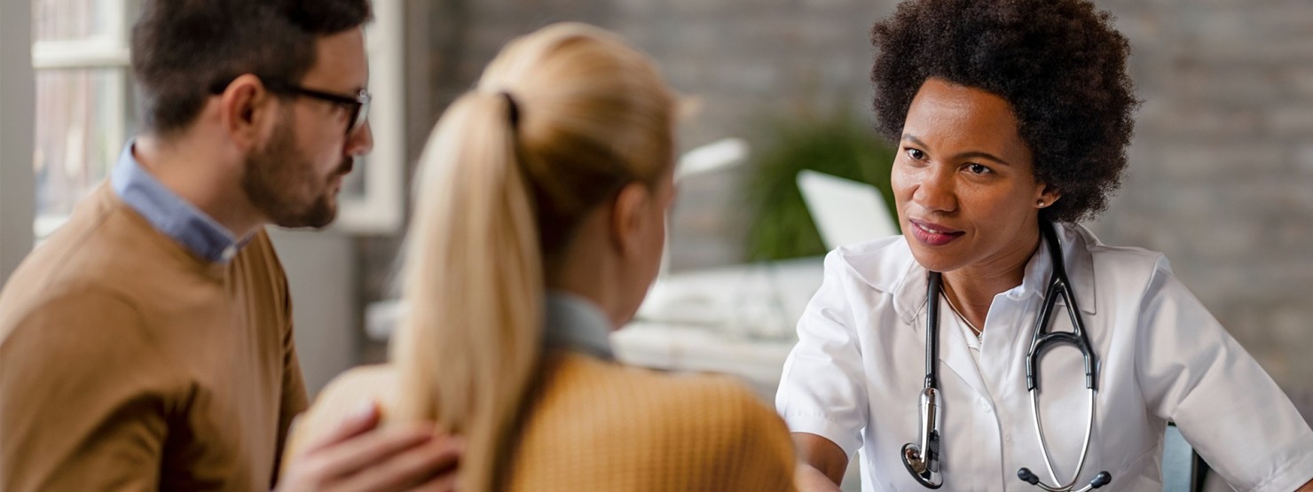 Ein etwa 45-jähriges Paar sitzt in einer Praxis und wird von einer etwa 35-jährigen Ärztin beraten.