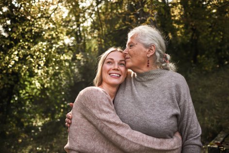 Eine etwa 25-jährige Frau und ihre etwa 75-jährige Großmutter stehen gemeinsam draußen im Grünen. Die junge Frau hat die Arme um die Mitte ihrer Großmutter geschlungen und lächelt glücklich. Ihr Blick schweift in die Ferne. Die Großmutter berührt sanft mit der Seite ihres Gesichts die Stirn der jungen Frau und hat ihre Hand sanft auf ihren Rücken gelegt.