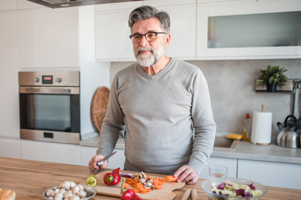 A smiling man with gray hair and beard is making a healthy lunch of vegetables at the kitchen counter