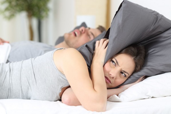 Eine Frau liegt mit ihrem Partner im Bett. Er liegt auf dem Rücken und schnarcht. Sie wirkt genervt und hält sich mit dem Kissen die Ohren zu.