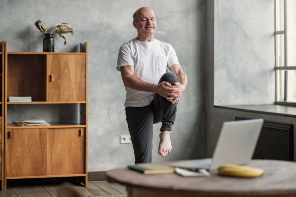Ein etwa 60 jähriger Mann betätigt sich sportlich in einem Wohnzimmer. Dabei dehnt er sein linkes Bein. Währenddessen schaut er auf einen Laptop, welcher auf einem Tisch steht.