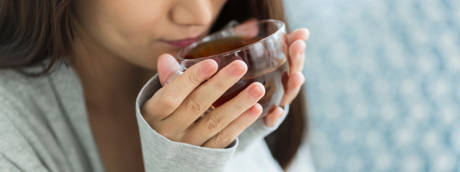 Eine etwa 30-jährige Frau hält eine Glastasse mit Tee zwischen beiden Händen und führt sie zum Mund.