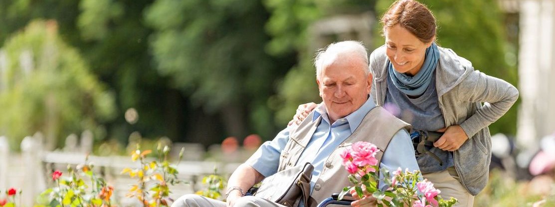 Ein Senior im Rollstuhl wird von einer Pflegerin entlang eines Rosenbeets geschoben.