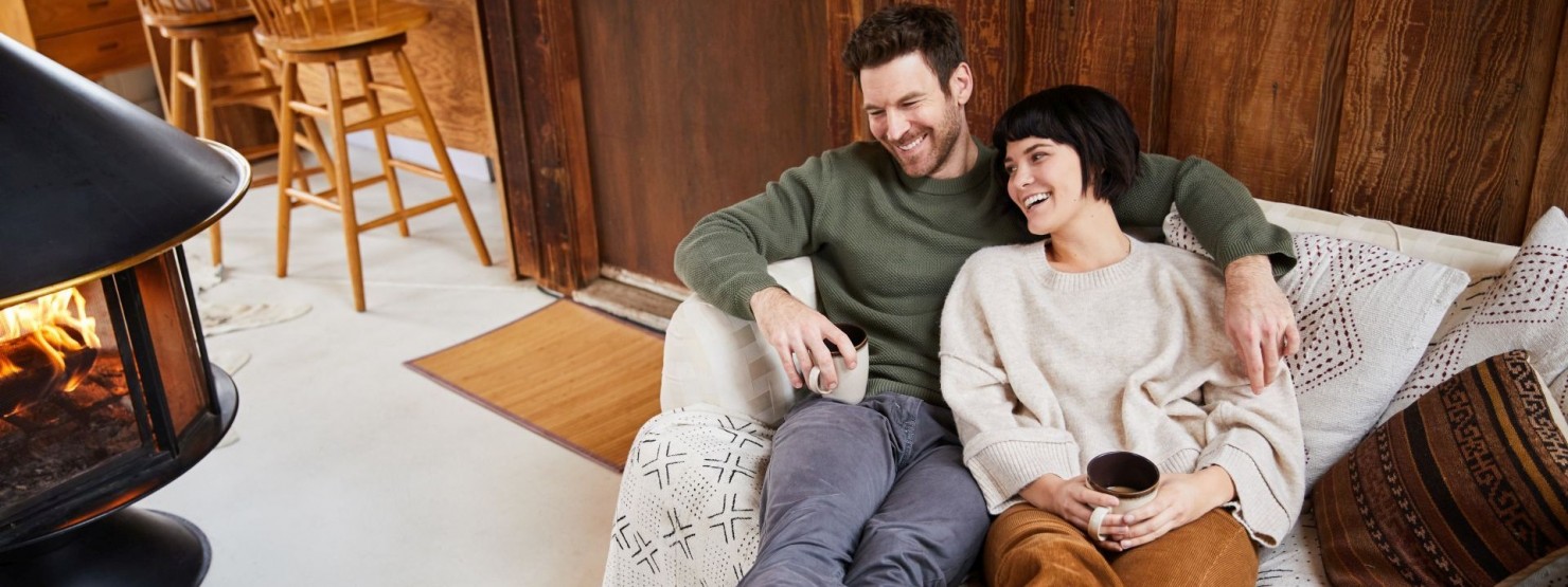 Ein Mann und eine Frau liegen gemeinsam aneinander gelehnt auf der Couch. Beide lachen und halten jeweils eine Tasse Kaffee in den Händen.