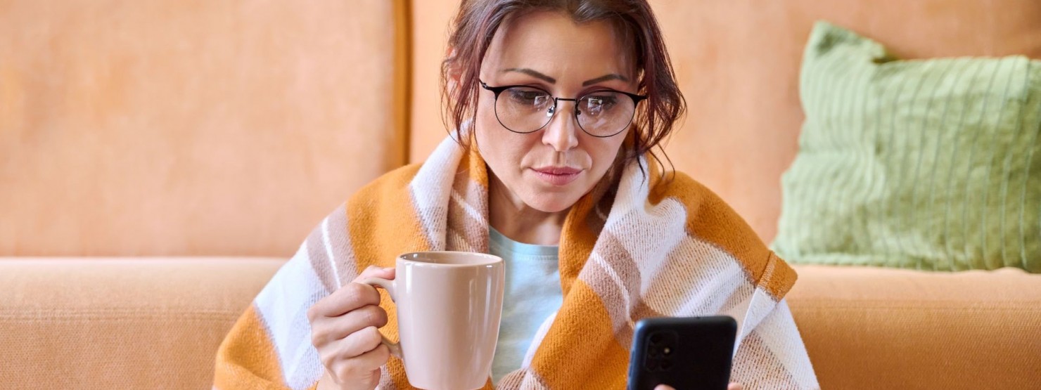 Eine Frau sitzt vor einem Sofa und schaut auf ihr Handy. Sie ist krank und hält ein Tasse in ihrer Hand.
