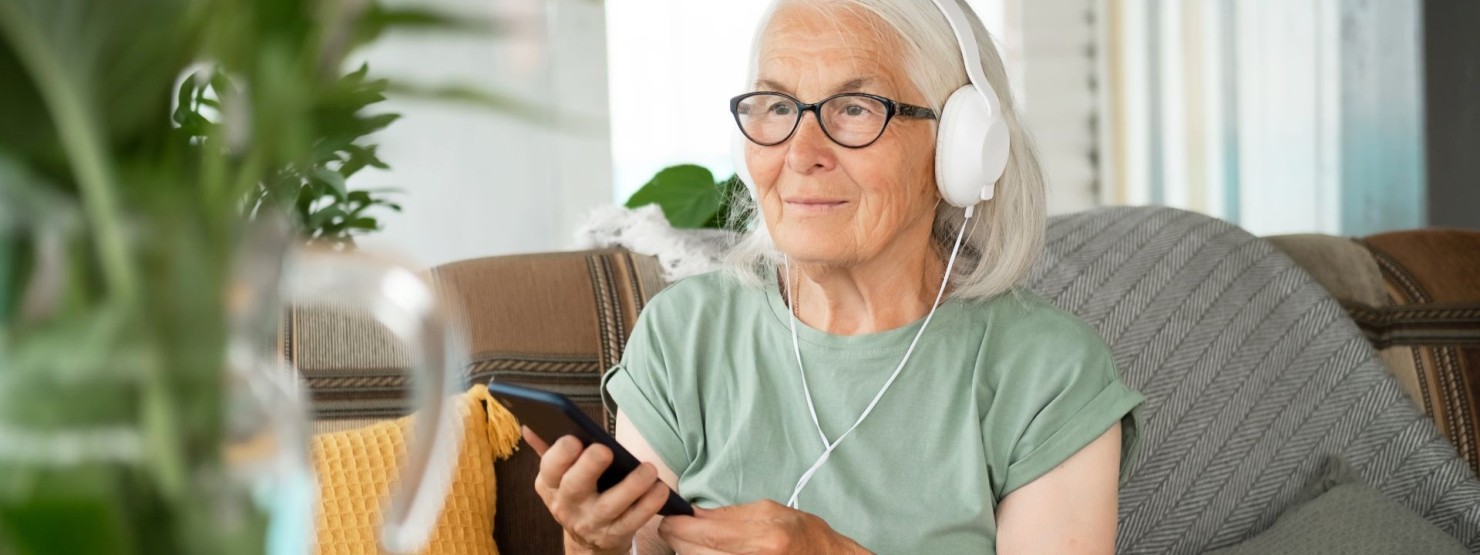 Eine Seniorin sitzt auf einem Sofa und hört über Kopfhörer einen Podcast.