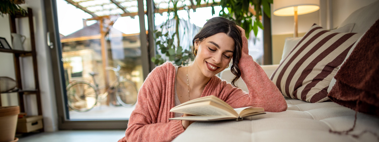 Eine etwa 30-jährige Frau liest mit lächelndem Gesicht ein Buch auf der Couch. Ihr Kopf liegt entspannt auf der Rückenlehne.
